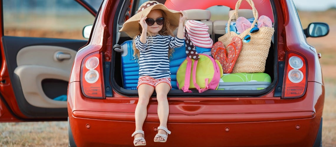 Une petite fille est assise sur le bord du coffre d'une voiture chargée de ses bagages et affaires de plage